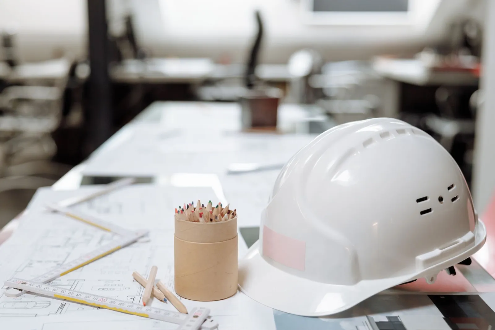 Close-up of an office desk with a white helmet, colored pencils, and architectural plans.