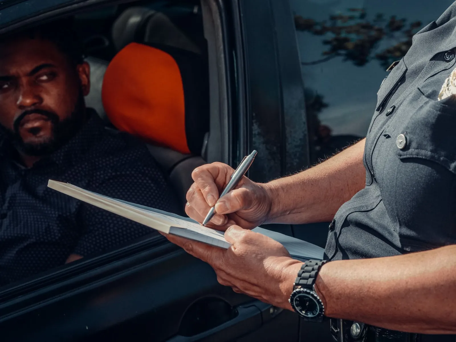 A police officer writes a ticket as the driver looks on from inside the car.