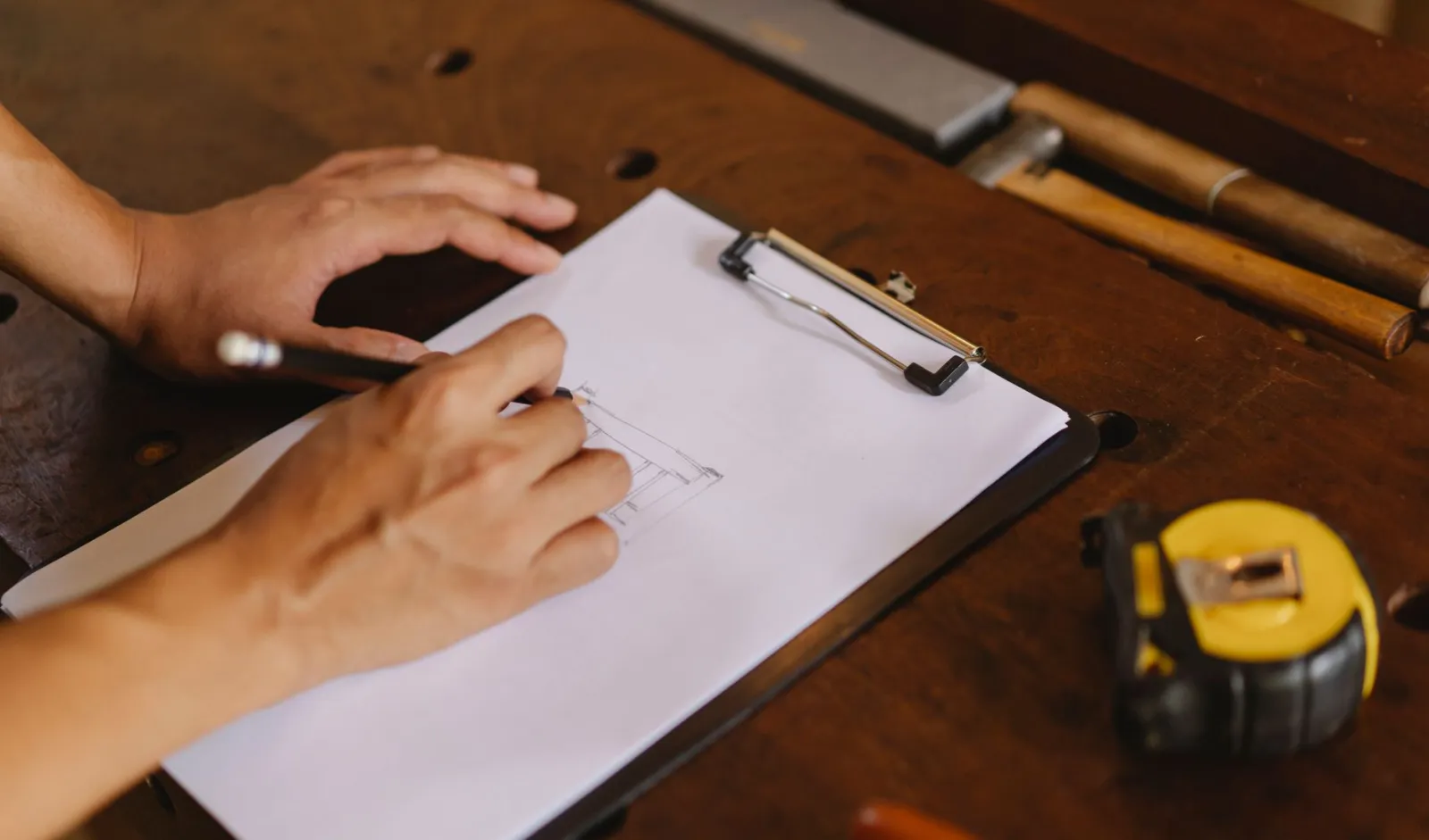 Unrecognizable male artisan making sketch of wooden product while standing at table with measuring tape and tools in professional studio